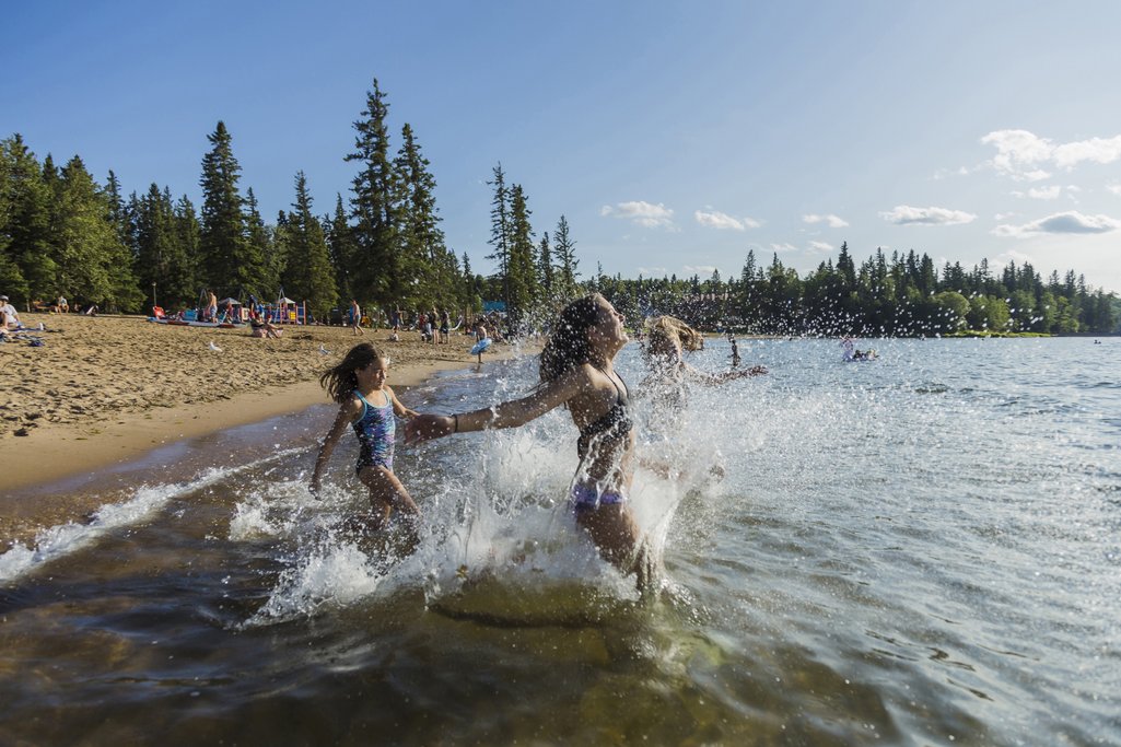 Two kids running into a lake from the beach and making a big splash with a forest surround the beach and lake