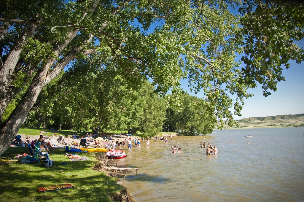 A bunch of people at a beach playing in the sand and water at Lake Deifenbaker on a blue sky summer day.