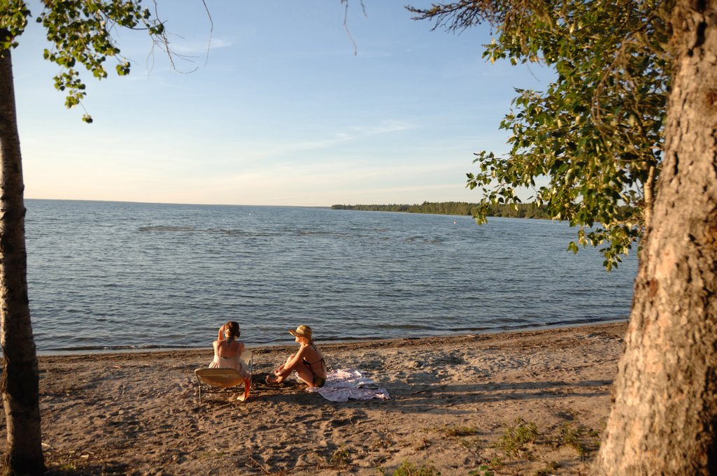 Two people sitting on a beach at Candle Lake Provincial Park at golden hour on a calm summer evening.