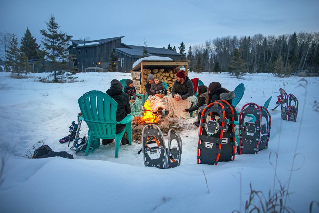 a family sitting around a campfire outside during winter with their snowshoes standing in the snow beside them and Waters Edge Eco Lodge in the background
