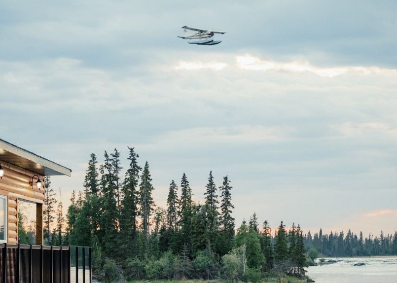 airplane flying over an island on water by wooden building, trees and mountains in distance